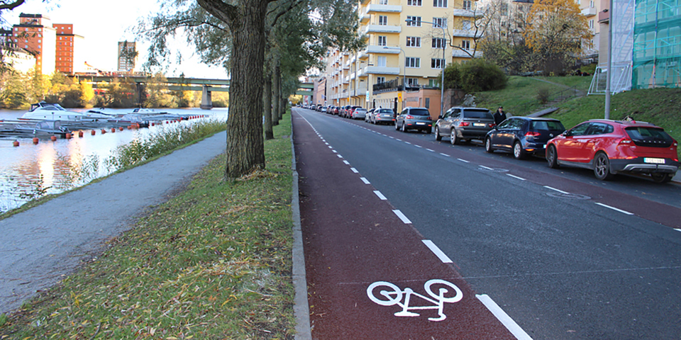 Cykelväg, bilväg och en gångväg av grus med träd och gräs runt om sig. Gångvägen ligger vid vattnet. Parkerade bilar och hus. Foto.